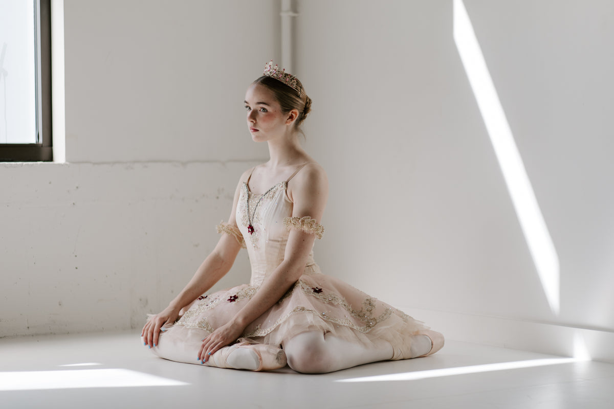 A ballet dancer in a pale tutu and tiara sitting on the floor of a sunlit studio